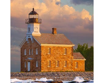 May include: A stone lighthouse with a white tower and a black lantern on a rocky coast. The lighthouse is surrounded by blue water and white waves. The sky is a soft pink and orange.