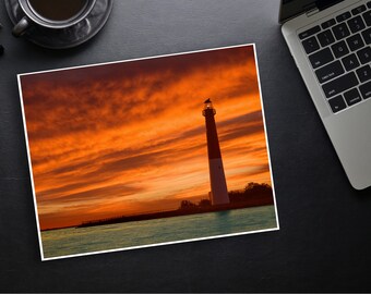 May include: A tall white lighthouse stands against a vibrant orange and red sunset sky. The lighthouse is silhouetted against the clouds, with the sun setting behind it. The water in the foreground is calm and reflects the colors of the sky.