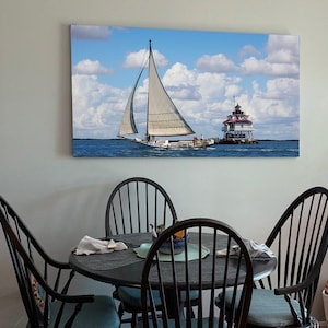 May include: A framed print of a sailboat sailing past a lighthouse on a sunny day with a blue sky and white clouds. The print is hanging on a wall above a black dining table with four chairs.