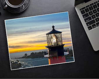 May include: A photograph of a lighthouse at sunset. The lighthouse is red and black with a glass lantern room. The sky is a mix of blue, orange, and yellow. The image is on a black surface with a laptop and coffee cup.