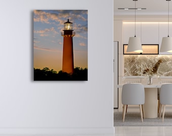 May include: A vertical canvas print of a lighthouse at dusk. The tall, orange lighthouse stands against a sky of blue and orange hues, with fluffy clouds. The top of the lighthouse is lit, and the bottom is obscured by dark trees.