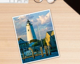 May include: A white lighthouse with a black roof stands tall against a blue sky with white clouds. The lighthouse is surrounded by water and a small dock with wooden pilings. There are several boats docked at the pier.