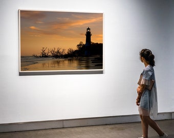 May include: A framed photograph of a lighthouse silhouetted against a sunset sky. The lighthouse is on a beach with a few trees in the foreground. A person is standing in front of the photograph, looking at it.