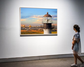 May include: A framed photograph of a lighthouse with a white and brown exterior, a glass lantern, and a metal railing. The lighthouse is situated on a grassy field with a city skyline in the background. The sky is blue with white clouds. A person is standing in front of the photograph, looking at it.
