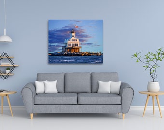 May include: A framed canvas print of a white lighthouse with a red, white, and blue American flag waving on top. The lighthouse sits on a rocky island surrounded by blue water under a cloudy sky. The print is displayed above a gray sofa.