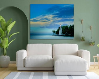 May include: A white lighthouse with a red roof stands on a rocky shore, surrounded by trees. The lighthouse is illuminated, casting a warm glow against the blue sky and calm water. The sky is a vibrant blue with wispy clouds.
