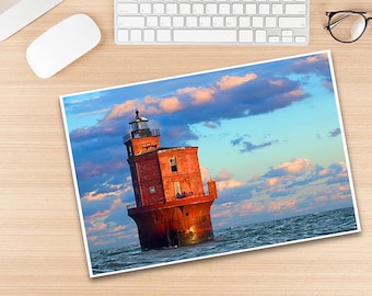 May include: A photograph of a red brick lighthouse in the ocean under a cloudy sky. The lighthouse has a black top and is surrounded by water. The image is on a wooden desk with a keyboard, mouse, and glasses.