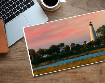 May include: A white lighthouse stands tall against a pink and orange sunset sky. The lighthouse is surrounded by trees and a body of water.