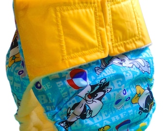 May include: A cloth diaper featuring a vibrant blue background with cartoon characters. The diaper has a yellow velcro closure and a soft, absorbent interior. The design includes images of Sylvester the Cat, Bugs Bunny, and other cartoon characters.