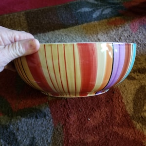 May include: A large, colorful ceramic bowl with a striped pattern in shades of yellow, orange, red, purple, and blue. The bowl is perfect for serving salads, fruit, or snacks.