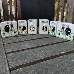 May include: Six white ceramic spice jars with rooster illustrations and black lettering. The jars are labeled "Salt", "Allspice", "Paprika", "Ginger", "Thyme", and "Pepper".
