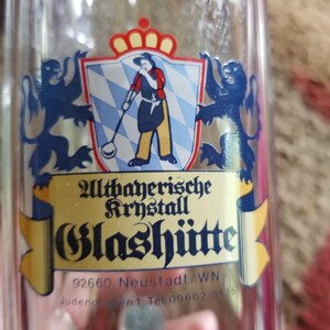 May include: A clear glass bottle with a label featuring a crest with a figure holding a tool, a crown, and two blue lions. The label also includes the text "Altbayerische Krystall Glashütte".