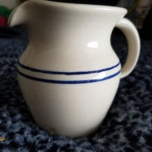 May include: A white ceramic mug with a black handle and the text "But First, Coffee" in a bold, black font. The mug is set against a blurred background, suggesting a cozy setting. The mug appears to be approximately 4 inches tall.