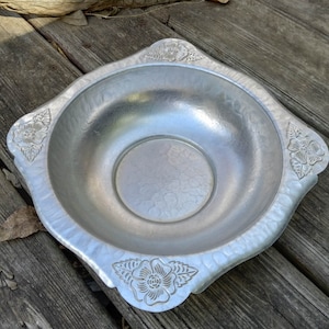 May include: A silver metal bowl with a square shape and a scalloped edge. The bowl has a hammered finish and is decorated with floral designs.