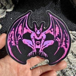 May include: A die-cut sticker featuring a stylised gargoyle creature with large, expansive wings. The design is rendered in a vibrant neon pink colour against a solid black background, showcasing sharp claws and pointed ears in a crouching pose.