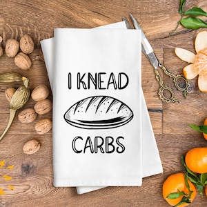 Könnte beinhalten: Ein weißes Küchentuch mit dem Text "I KNEAD CARBS" in schwarzen Buchstaben. Eine Schwarzweißillustration eines Brotlaibs befindet sich über dem Text.