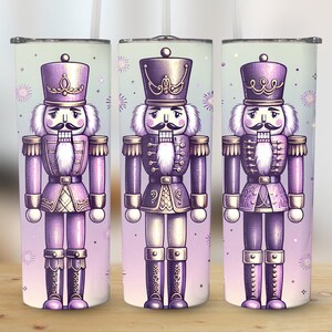 May include: Three stainless steel tumblers with a festive nutcracker design. Each tumbler features a purple and gold nutcracker illustration with a white mustache and a decorative hat. The background is a light purple with snowflake accents.