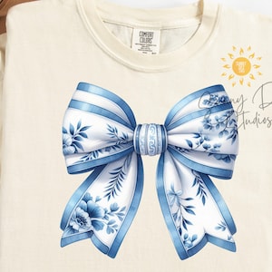 May include: A blue and white bow with a floral pattern on a cream colored t-shirt. The bow is tied in a knot and the ends are flowing. The t-shirt is a plain, light cream color.