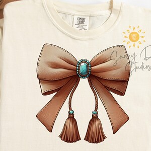 May include: A brown bow with turquoise gem embellishment and tassels. The bow is on a cream coloured t-shirt with the text "Sunny Day Studios" in black.