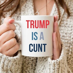 May include: White ceramic mug with the text "TRUMP IS A CUNT" in red, blue, and navy blue. The mug is held by a person wearing a cream-colored sweater.