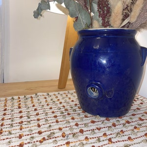 May include: A cobalt blue ceramic vase with a round opening and two small handles. The vase holds dried flowers and greenery. The vase sits on a wooden surface next to a patterned runner.