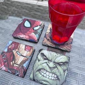 K&ouml;nnte beinhalten: Vier quadratische Schieferuntersetzer mit Marvel-Comicfiguren: Spider-Man, Iron Man, Hulk und Thanos. Ein rotes Plastikglas steht auf einem der Untersetzer. Die Untersetzer befinden sich auf einer grauen, strukturierten Oberfl&auml;che.