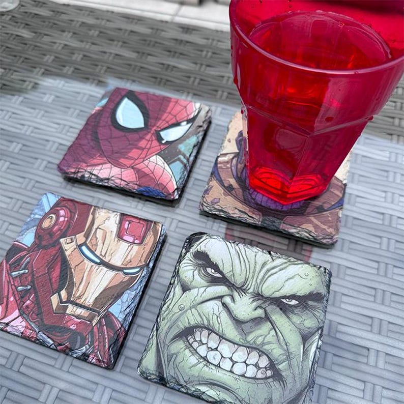 K&ouml;nnte beinhalten: Vier quadratische Schieferuntersetzer mit Marvel-Comicfiguren: Spider-Man, Iron Man, Hulk und Thanos. Ein rotes Plastikglas steht auf einem der Untersetzer. Die Untersetzer befinden sich auf einer grauen, strukturierten Oberfl&auml;che.
