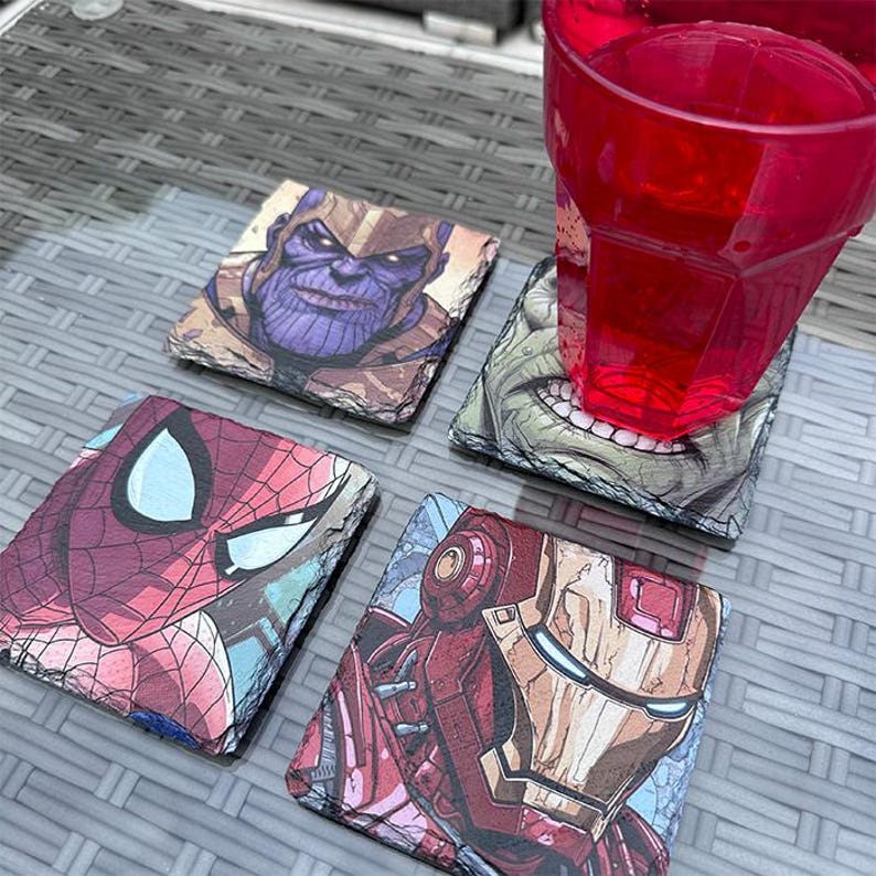 K&ouml;nnte beinhalten: Vier quadratische Schieferuntersetzer mit Marvel-Comicfiguren: Thanos, Hulk, Spider-Man und Iron Man. Ein rotes Plastikglas steht auf dem Hulk-Untersetzer. Die Untersetzer haben ein nat&uuml;rliches, strukturiertes Aussehen mit farbenfrohen Charakterillustrationen.
