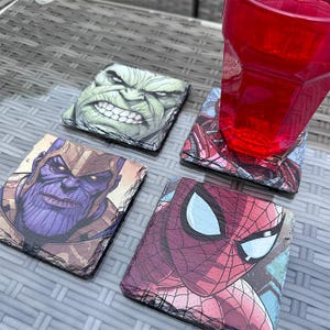K&ouml;nnte beinhalten: Vier quadratische Schieferuntersetzer mit Marvel-Comicfiguren: Hulk, Thanos, Spider-Man und Iron Man. Die Untersetzer befinden sich auf einer grauen, gewebten Oberfl&auml;che, mit einem teilweise sichtbaren roten Glas.