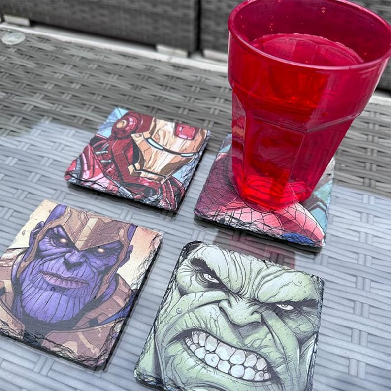 K&ouml;nnte beinhalten: Vier quadratische Schieferuntersetzer mit Marvel-Comicfiguren: Iron Man, Spider-Man, Thanos und Hulk. Ein roter Plastikbecher mit Wasser steht auf einem der Untersetzer. Die Untersetzer stehen auf einem Glastisch.