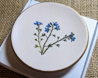 Puede incluir: Un posavasos redondo de cerámica blanquecina con borde dorado, con un delicado arreglo de flores azules nomeolvides. El posavasos descansa sobre una caja blanca, sobre un fondo beige texturizado.