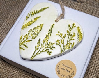 May include: A white ceramic heart-shaped ornament with a green floral design. The ornament is hanging from a twine string and has a brown paper sticker that says "handmade with love".