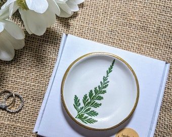 May include: A white ceramic ring dish with a gold rim and a green fern design. The dish is on a white background and has a brown paper tag that says "handmade with love".