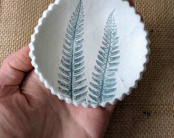 May include: A small, white ceramic dish with a scalloped edge. Two blue-green fern leaf designs are printed on the inside of the dish. The dish is round and held in a hand.