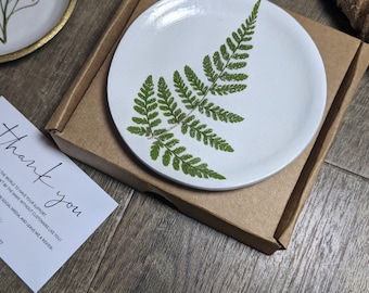 May include: A white ceramic trinket dish with a pressed fern design. The dish is packaged in a brown cardboard box with a thank you note.