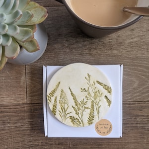 Könnte beinhalten: Ein runder, weißer Untersetzer aus Keramik mit einem grünen Blumenmuster. Der Untersetzer hat einen braunen Papieretikett mit dem Text "handgefertigt mit Liebe" und einem kleinen Herzen.
