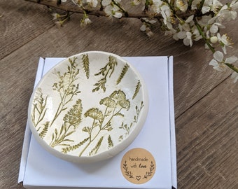 Puede incluir: Un pequeño plato de cerámica blanca con un diseño floral verde. El plato está sobre una caja blanca con una pegatina redonda marrón que dice "hecho a mano con amor".