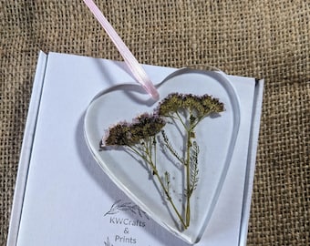 May include: A clear heart-shaped resin ornament with a pressed dried flower arrangement inside. The flower is a light purple and green color. The ornament has a pink ribbon attached to the top. The ornament is on a white background with the text "KWCrafts & Prints" printed on the bottom.