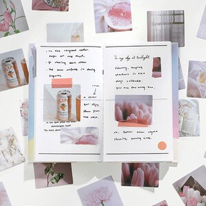 May include: A collection of square stickers featuring various images, including flowers, fruit, and a can of soda. The stickers are arranged around an open notebook with handwritten text and a few more stickers on the pages. The notebook has a rainbow gradient on the cover.
