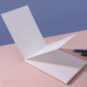 May include: A white sketchbook with a soft cover is open to reveal blank pages. Two coloured pencils are lying on a pink surface next to the sketchbook.