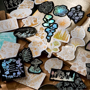 May include: Assortment of gold and iridescent holographic stickers featuring floral, nature, and geometric designs. The stickers are on a brown surface with a book in the background.