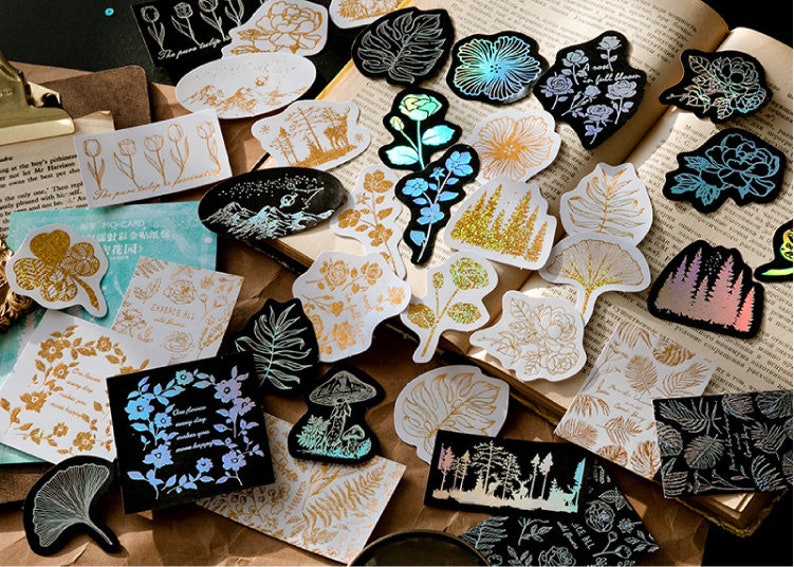 May include: Assortment of gold and iridescent holographic stickers featuring floral, nature, and geometric designs. The stickers are on a brown surface with a book in the background.