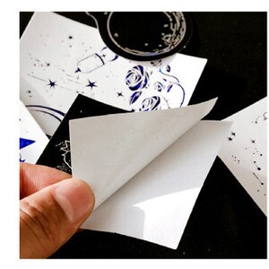 May include: A set of holographic stickers featuring celestial and floral designs. The stickers are square and have a white backing. The designs include a crescent moon, stars, a rocket ship, and roses.