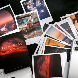 May include: A stack of instant photos with various subjects, including a sunset, fireworks, and a restaurant interior. A small book titled "Castle in the Sky" is visible in the foreground. The book has a red and orange sunset on the cover with the text "Maybe you've walked into someone else's landscape".