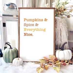 Puede incluir: Una impresión enmarcada con el texto "Pumpkins & Spice & Everything Nice." en texto naranja, amarillo y rojo. La impresión está sobre una superficie blanca y esponjosa con una calabaza blanca y una calabaza verde en primer plano.