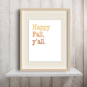 Puede incluir: Una impresión enmarcada con el texto "Happy Fall, y'all." en un degradado de colores naranja y amarillo. El marco es de madera clara y la impresión está sobre un fondo blanco.