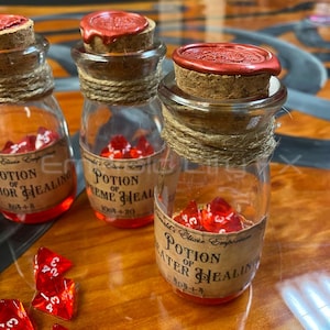 May include: Glass potion bottles with cork stoppers sealed with red wax. Each bottle is labeled with text and filled with red dice. The bottles are wrapped with twine. The labels read "Potion of Minor Healing", "Potion of Supreme Healing", and "Potion of Greater Healing".