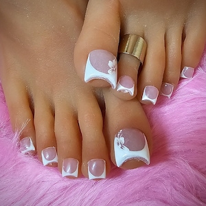 May include: Close-up of feet with white and pink square-shaped toenails with white flower designs. A gold toe ring is on the second toe.
