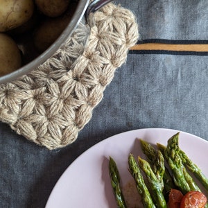 Könnte beinhalten: Eine rustikale Küchenszene mit einem Untersetzer aus Jute mit Sternmuster, einem Metalltopf mit Kartoffeln und einem rosa Teller mit Spargel und Tomaten. Die Szene ist auf einer grauen Tischdecke mit goldenem Streifen aufgebaut.