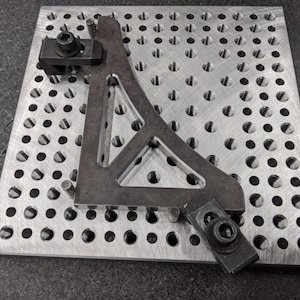 May include: A black metal triangle with a curved edge is clamped to a silver metal plate with a grid of holes. The triangle is secured with two black clamps.